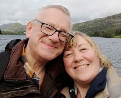 Smiling elderly man and woman enjoying a lakeside outdoor adventure in nature, with mountains and cloudy sky in the background.