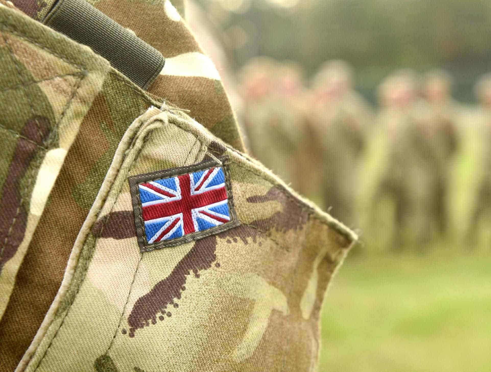 British military camouflage uniform with Union Jack patch on shoulder, outdoor setting with soldiers in the background.
