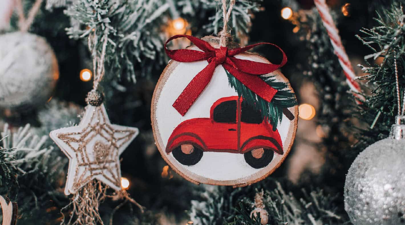 Hand-painted Christmas tree ornament featuring a red vintage scooter with green foliage, tied with a red ribbon, hanging on a decorated festive tree.