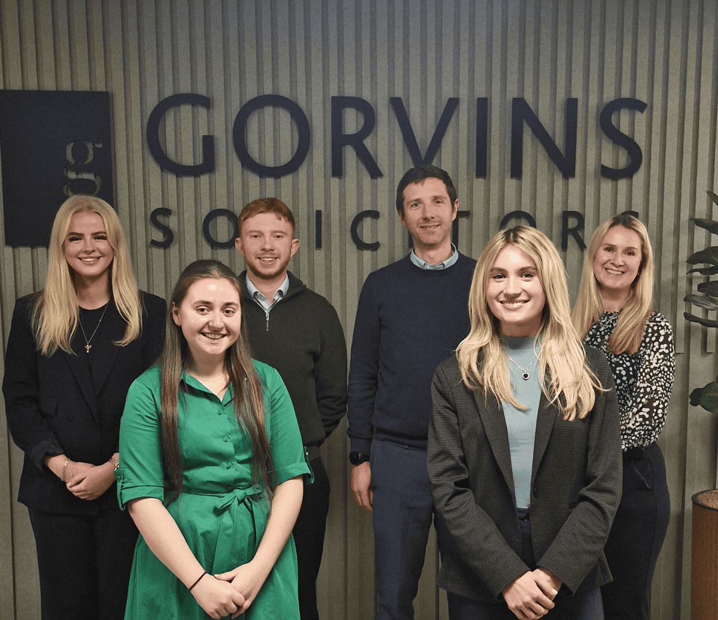 A group of six diverse professionals standing together in an office environment, smiling at the camera, with the Gorvins Solicitors logo visible on the wall behind them.