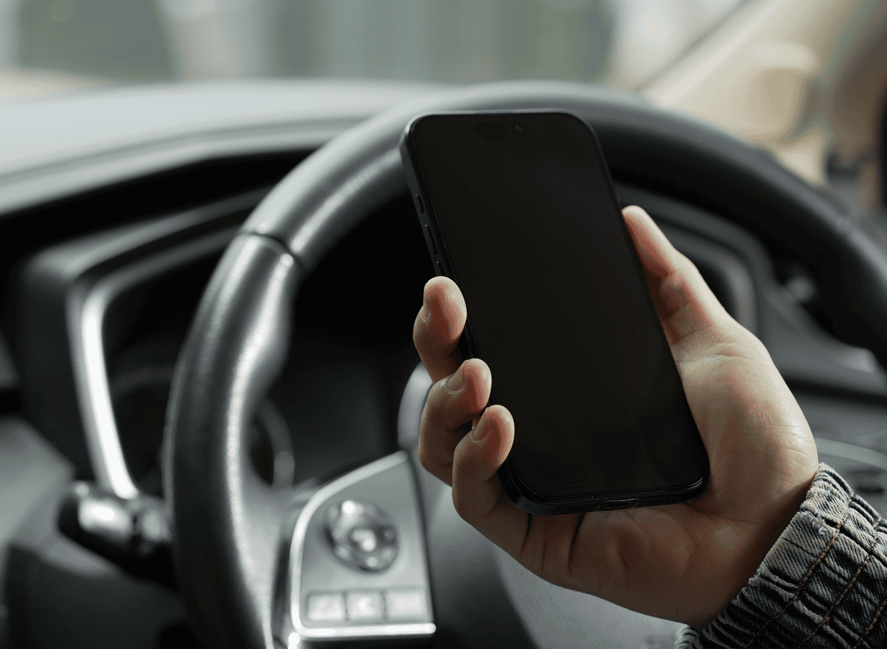 Person holding a smartphone while driving, with the steering wheel and dashboard visible in the background.