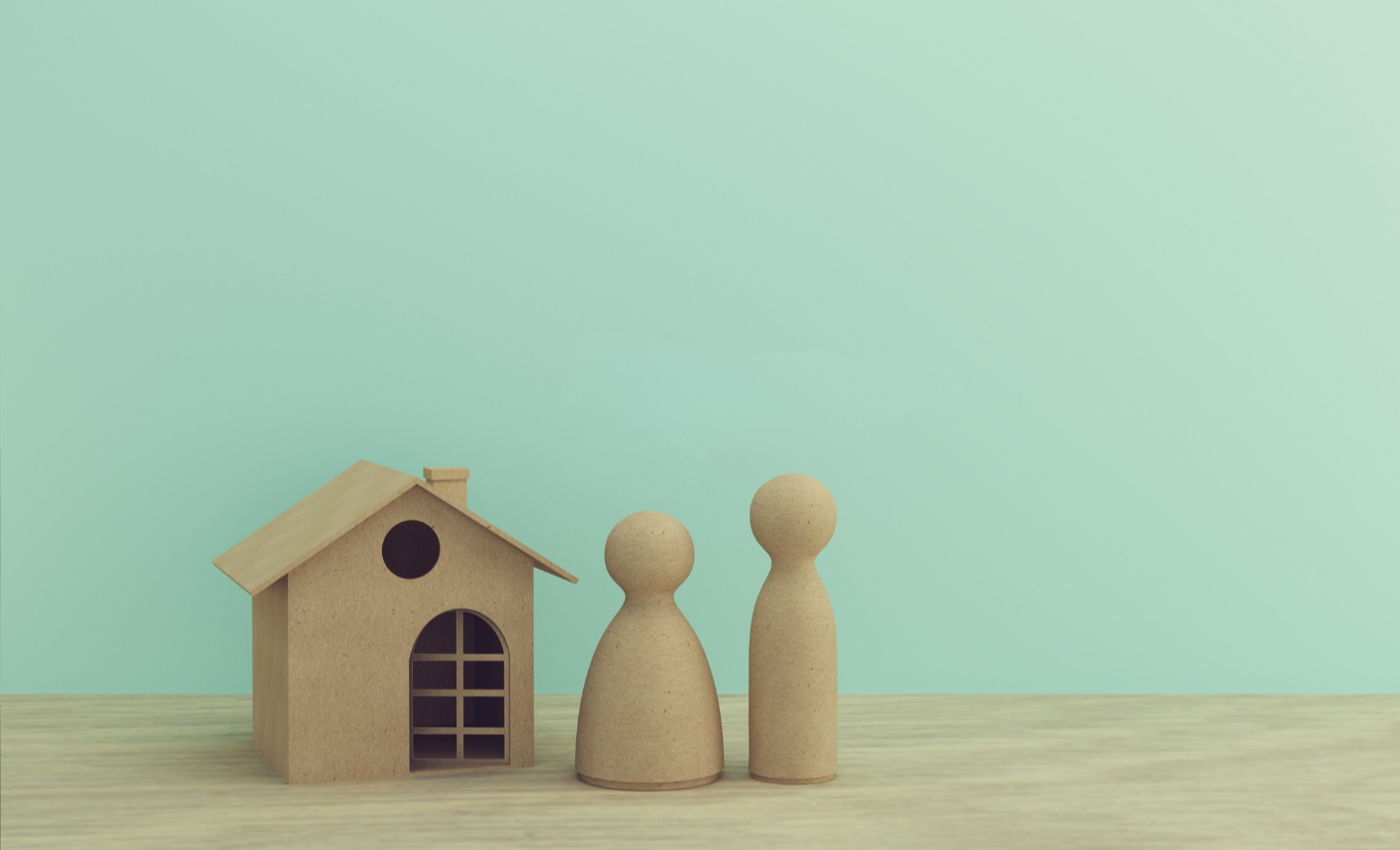 Simple wooden house model with two wooden figurines on a light wooden surface against a pastel green background.