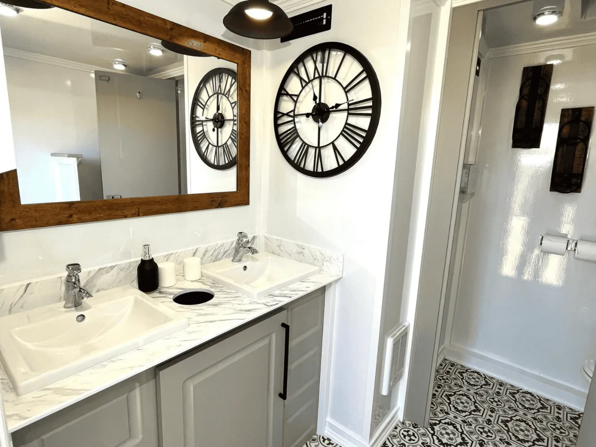 Inside a VannGo luxury restroom trailer: double vanity, marble counter, large mirror and wall clock