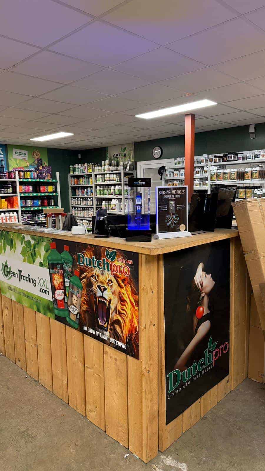 Eine Holztheke in einem Growshop mit Dutchpro- und Green Trading XXL-Bannern, einer Kasse, Werbeschildern und Regalen mit Flaschen und Gartenprodukten. An der Rückwand hängt eine große Uhr.