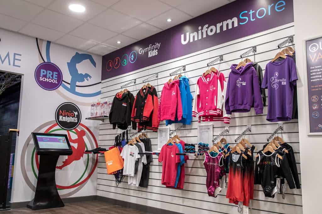 Brightly coloured kids' sports hoodies and uniforms displayed on hangers in a uniform shop with signage for Gymfinity Kids and Ninja Knights.