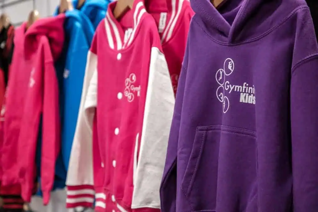 Brightly coloured kids' gymnastic hoodies and jackets displayed on hangers, featuring the Gymfin Kids logo, in a retail store. Perfect activewear for children engaging in sports activities.
