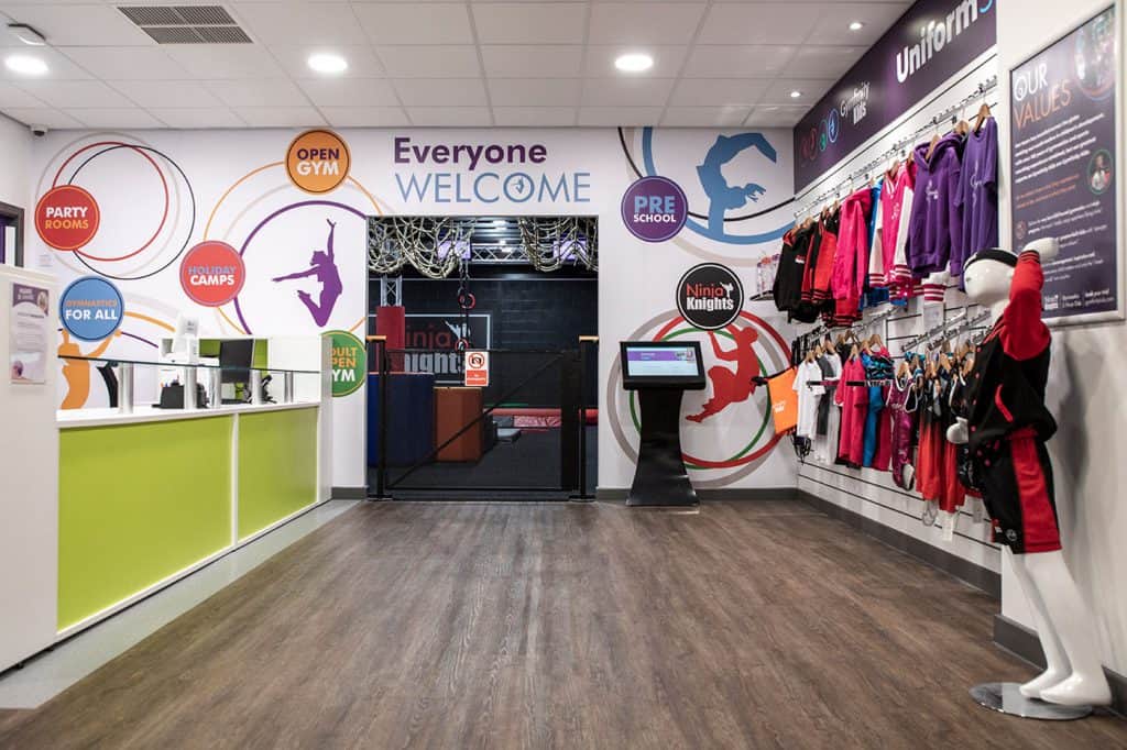 Bright sports centre reception area featuring colourful wall graphics, gym clothing for children, and an entrance to an indoor trampoline or ninja course zone. Designed to welcome families and promote activity.