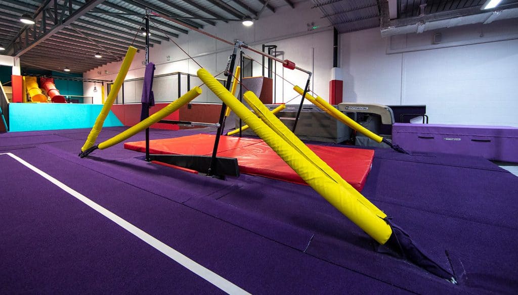 Flexible gymnastics training area with crash mats, poles, and equipment for aerial routines in a colourful indoor gymnasium.