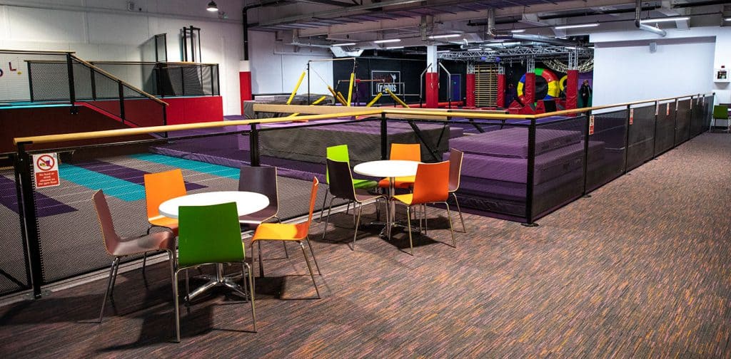 Brightly coloured chairs and round tables are placed near an indoor adventure play area with trampolines and climbing equipment in a spacious indoor soft play centre.