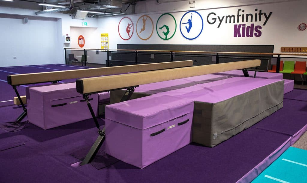 Springboard for gymnasts in a kids' gymnastics training area with purple floor mats and balance beams. The vibrant fun zone is designed for children's gymnastics practice and activities.