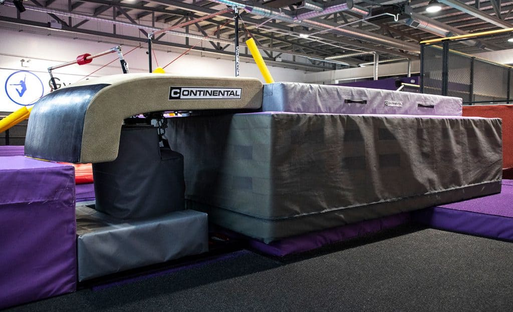 Flexible gymnastic vaulting table with padding and support structure, used for training and competitions in an indoor sports facility. The table is branded with "Continental" and is surrounded by mats for safety.