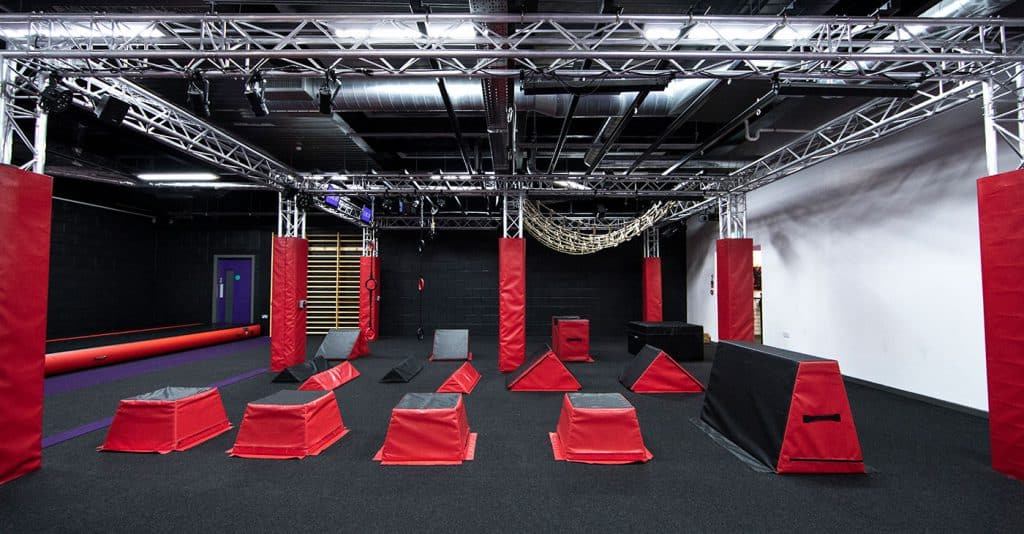 Climbing gym obstacle course with red padded equipment, ropes, and pull-up bars in an indoor training facility.