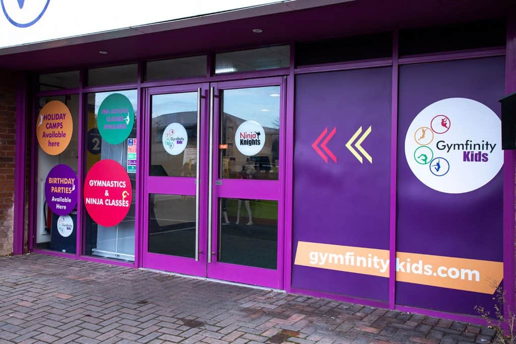 Colourful storefront windows of Gymfinity Kids advertising gymnastics, ninja classes, holiday camps, birthday parties, and pre-school classes in vibrant circles and signage.