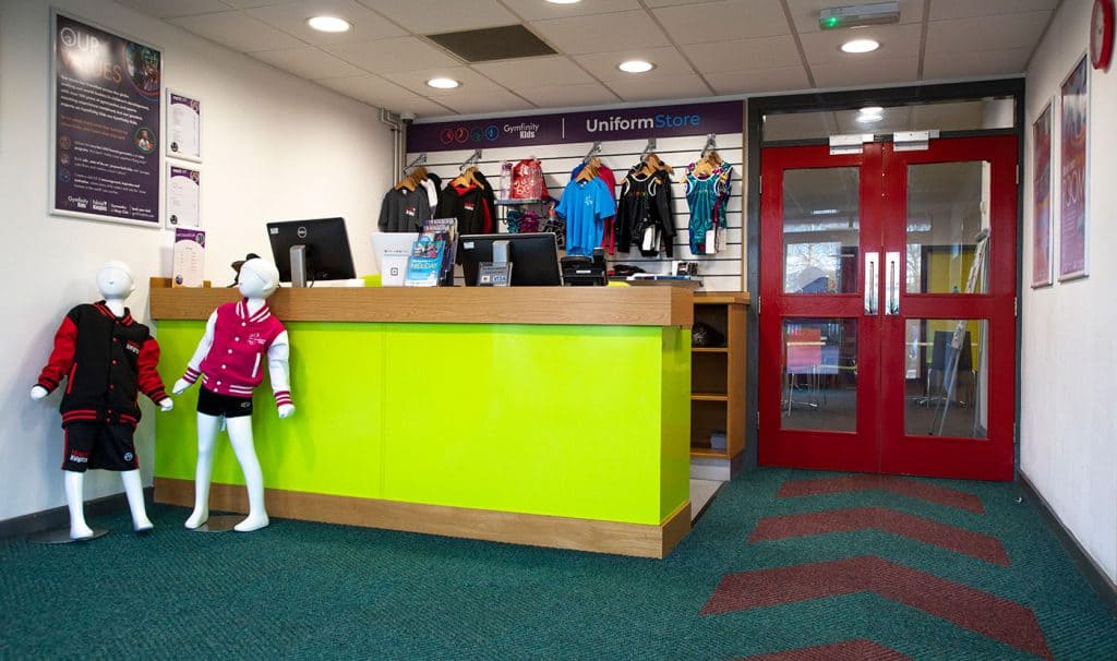 Brightly coloured sports merchandise store with notice board, mannequins in athletic clothing, and a checkout counter, located inside a fitness or sports centre.