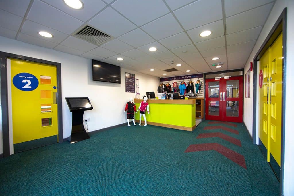 Bright and colourful indoor sports centre reception area with a sports uniform shop, mannequins dressed in sportswear, and vibrant yellow doors for party rooms.