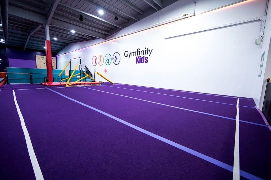 Race track flooring at Gymfinity Kids indoor play centre in purple with fitness equipment, colourful wall logos, and climbing wall in background, suitable for children's activities and exercises.