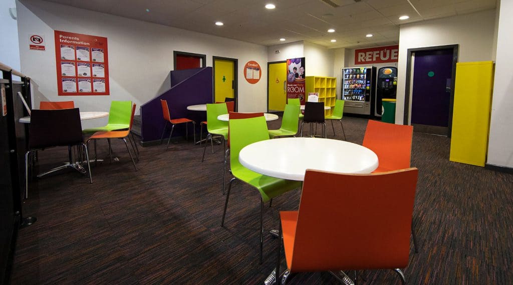 Brightly coloured seating area with orange, green, and black chairs around white round tables in a communal lounge space. There are vending machines, a refuge sign, and notice boards on the walls.