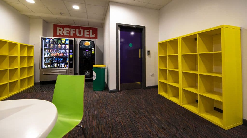 Vending machines and empty yellow shelving units in a brightly lit break room with a green chair and a table.