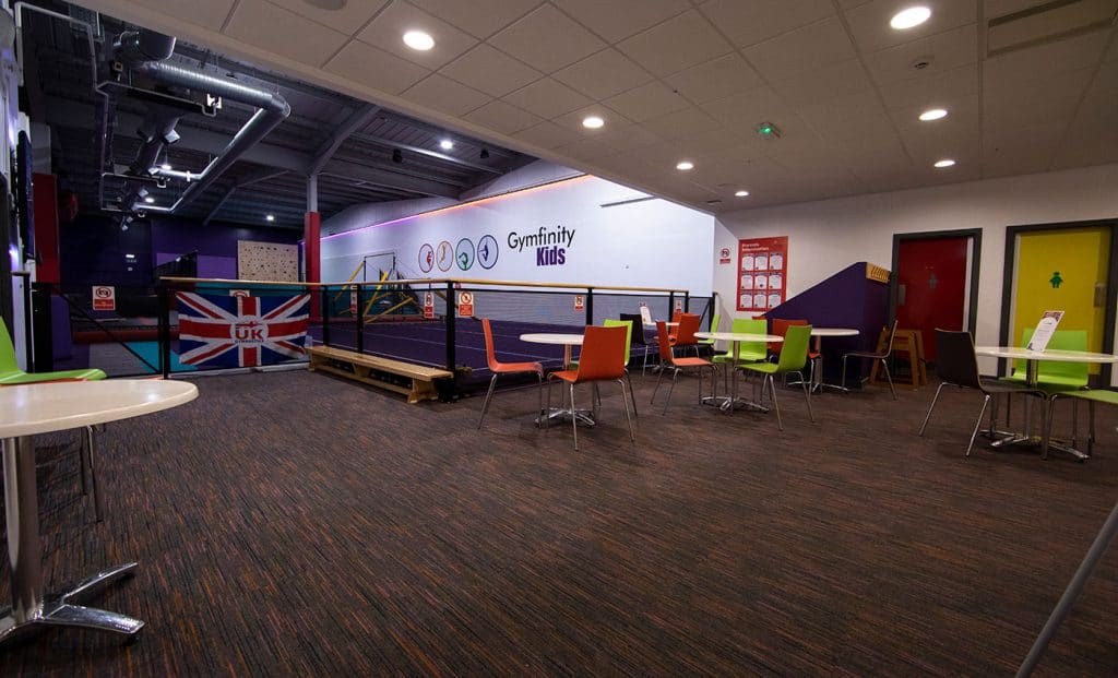 Bright indoor kids' gym area with seating, colourful chairs, and a football goal, featuring a sign that reads "Gymfinity Kids" and a British flag barrier, perfect for children's active play and activities.