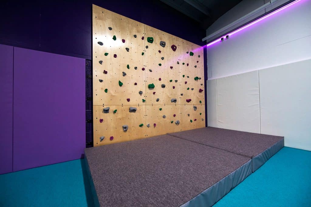 Climbing wall with colourful handholds in an indoor recreational facility, featuring padded flooring for safety, illuminated by purple LED strip lighting along the ceiling edge.