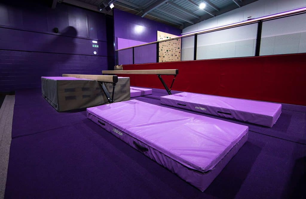 Floor vault gymnastics training area, purple mats and vaulting table, indoor sports facility, safety equipment, athletic training environment, vibrant lighting, purple and red wall accents, sports arena decor.