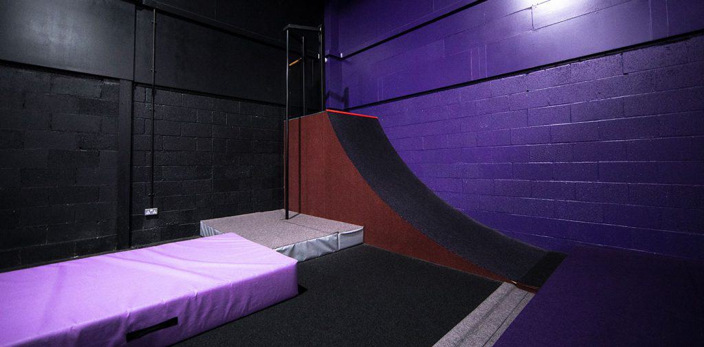 Ramp and padded landing zone in indoor skate park with black walls and purple lighting.