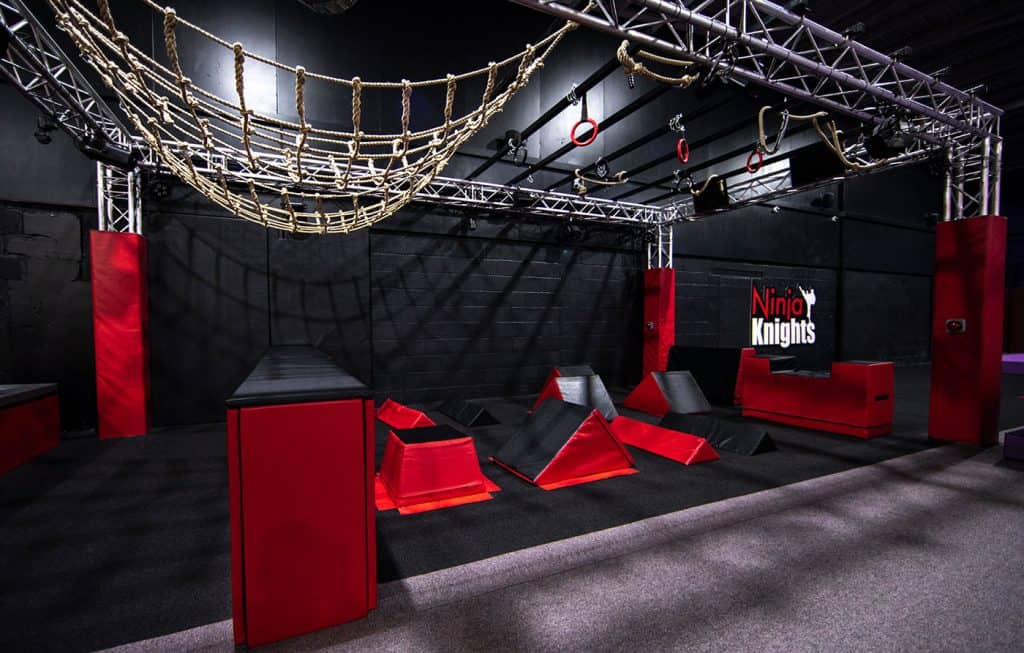 Climbing obstacle course with ropes, ramps, and hanging rings in an indoor ninja training facility, featuring a black and red colour scheme and branded "Ninja Knights" wall.