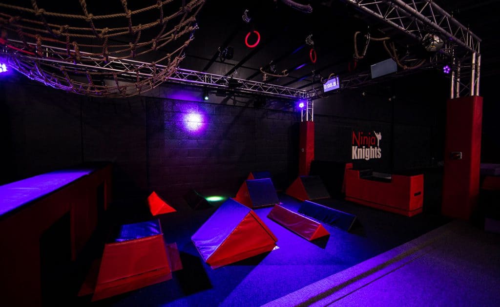 Obstacle course for kids with soft climbing shapes in an indoor adventure park, illuminated with colourful LED lights on a dark background.