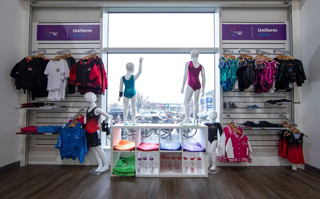 Brightly coloured kids' gymnastics uniforms and sportswear displayed on mannequins and racks inside a Gymfinity Kids store with large windows and parking outside.
