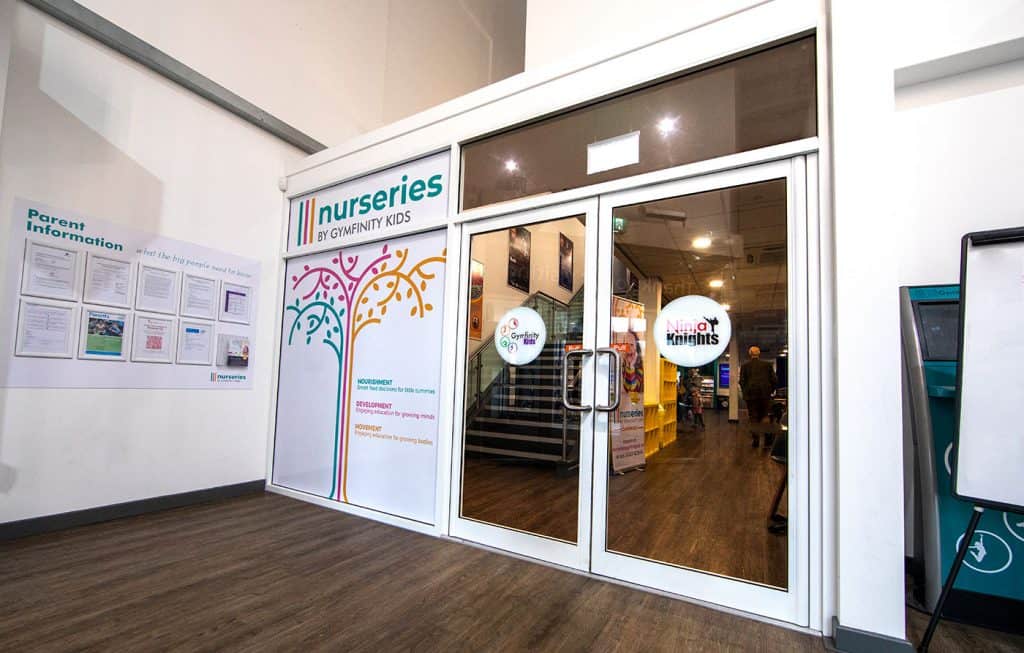 Nurseries entrance with glass doors, colourful signage, and parent information board inside modern facility, part of Gymfinity Kids childcare centre.