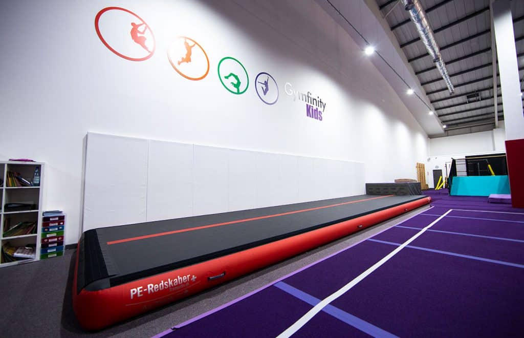 PE Redskaber trampoline on gymnasium floor in gymnastics training facility with colourful logo wall art and training equipment in background.
