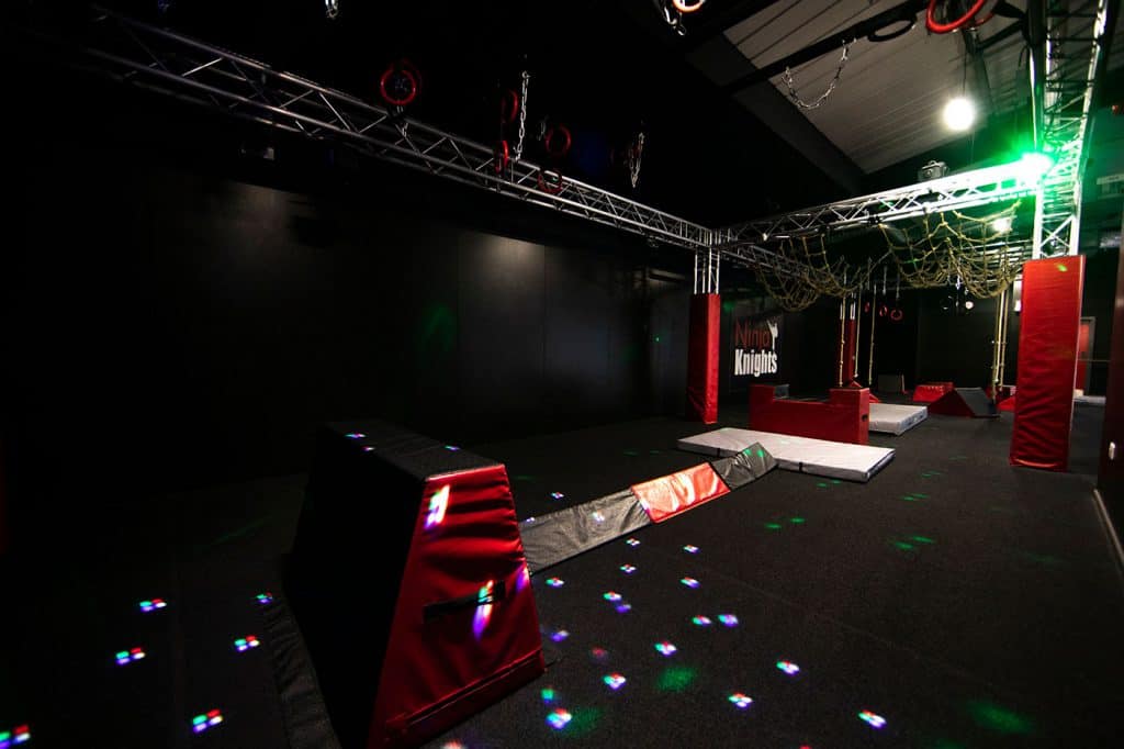 Climbing ninja training obstacle course with padded surfaces and hanging ropes in a dark indoor setting.