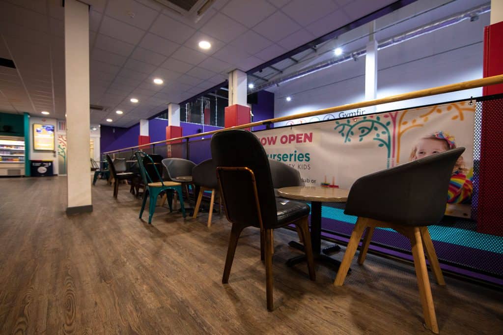 Light wood flooring with multiple chairs lining a partition in a modern indoor play area or cafe, featuring colourful decor and signage for nursery or kids' activities, well-lit with ceiling spotlights.