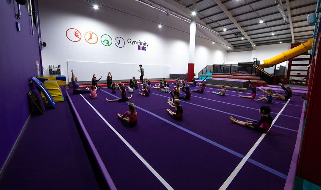 Kids participating in a gymnastics class at Gymfinity Kids indoor gym, doing stretches on purple mats under supervision in a bright, spacious facility with colourful equipment and a slide.