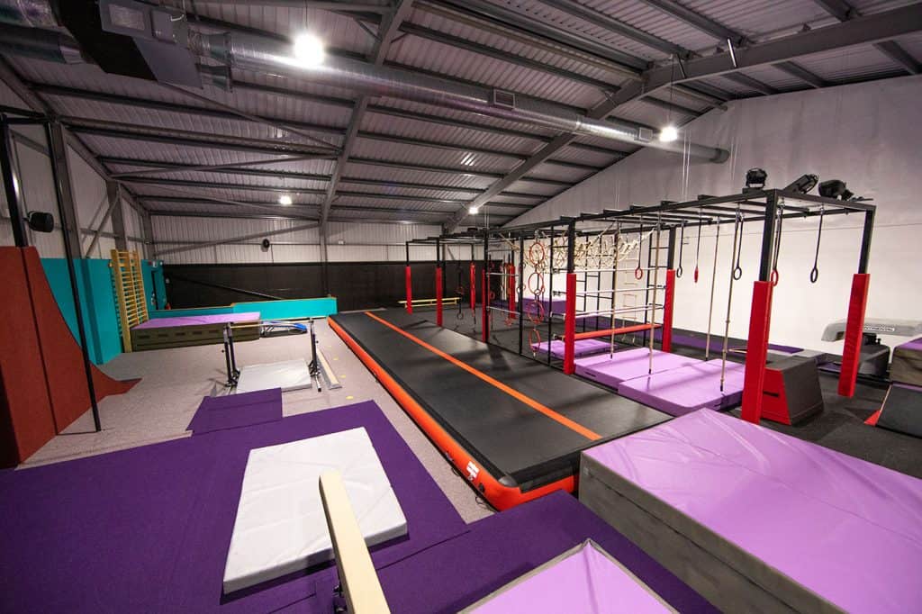 Climbing and gymnastic training area with padded mats, rings, monkey bars, and a trampoline inside a spacious indoor sports facility.