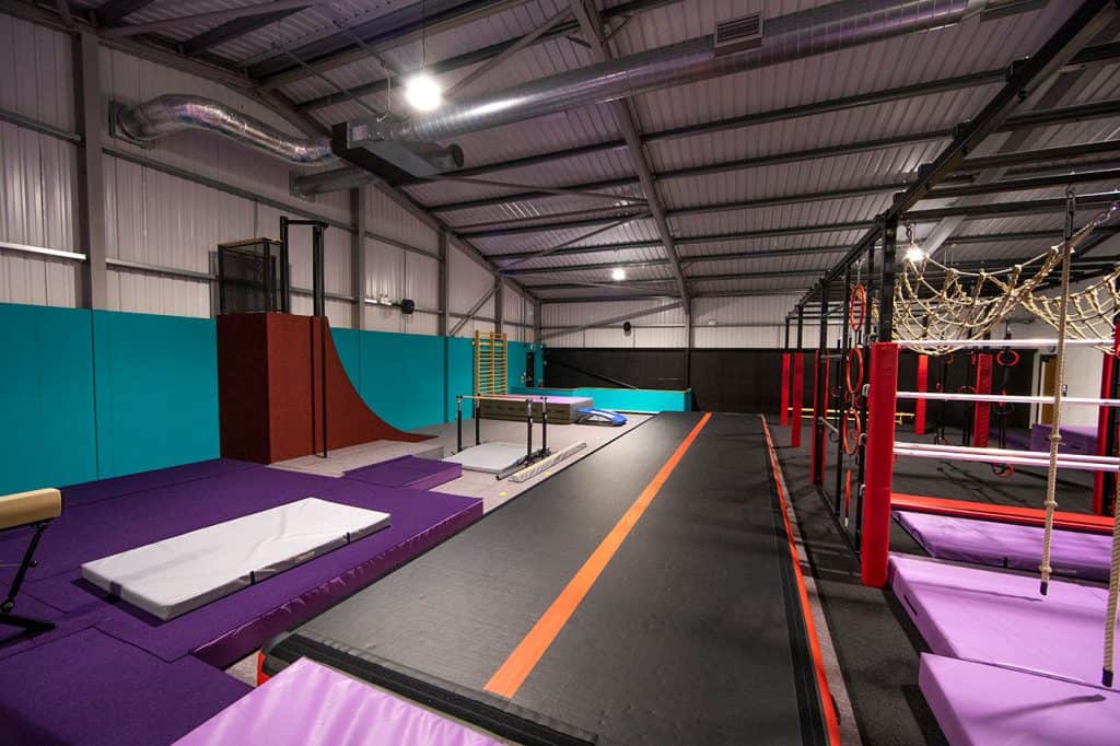 Climbing gym with padded floors, ropes, rings, and obstacle elements designed for indoor fitness and adventure training. Spacious industrial-style interior with colourful climbing and obstacle setups.