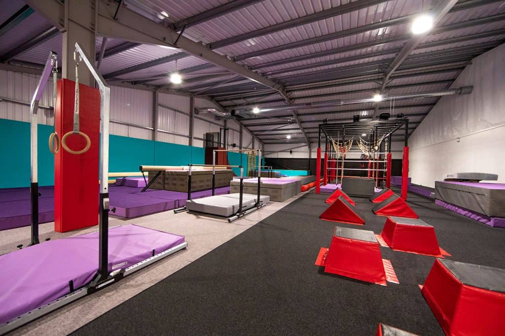 Climbing and obstacle course equipment inside an indoor gym, suitable for training and fitness activities. The space features padded mats, climbing frames, and red obstacles for agility exercises.