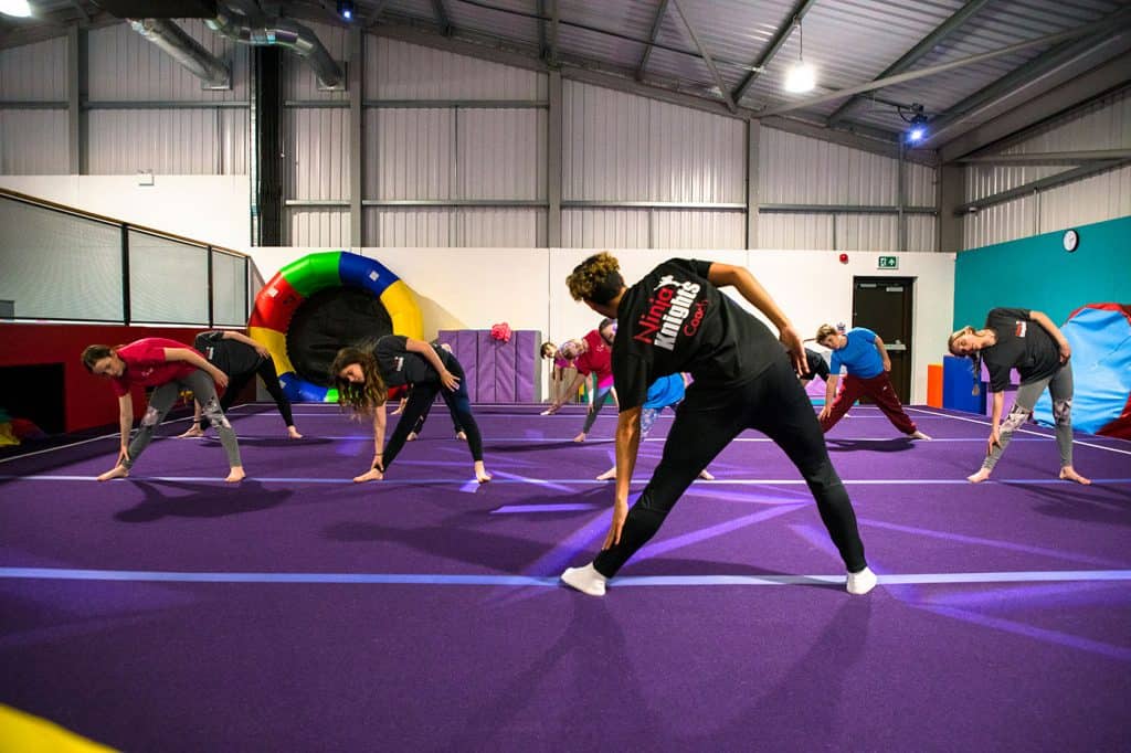 Legs stretch warm-up exercise at indoor gymnastics training session in a trampoline park, with instructor guiding a group of young gymnasts on a purple cushioned arena.