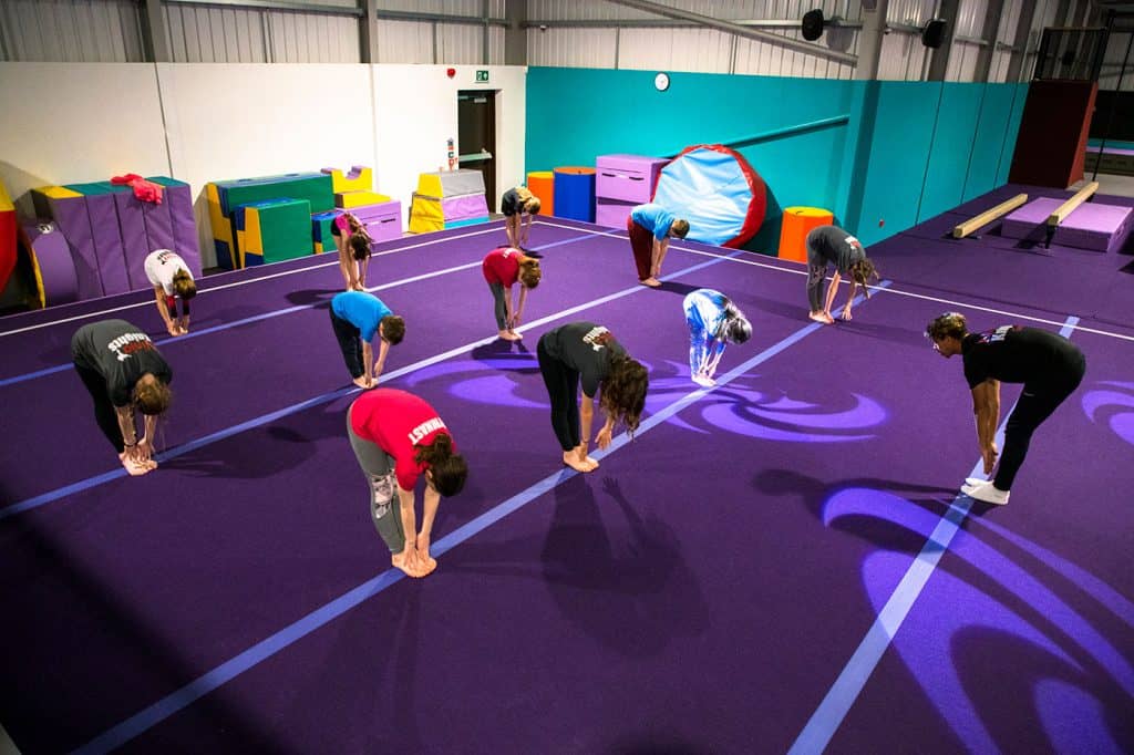 Boys and girls are practicing a gymnastics stretch on a purple mat in an indoor gymnasium with colourful equipment in the background.