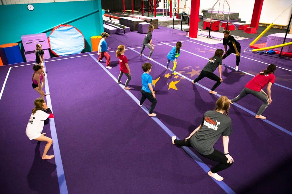 Jumping children participating in a gymnastics class at an indoor trampoline park, engaging in exercises on a large purple mat with stars design for fun and fitness.