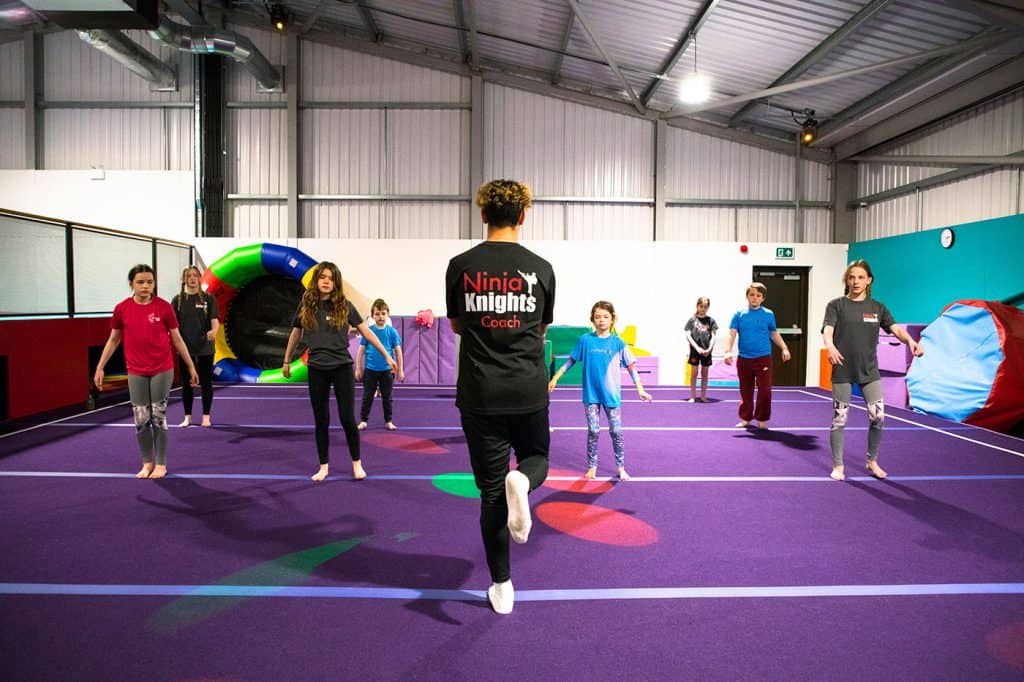 Jumping fitness class for kids in indoor trampoline park, instructor supervising children during exercise session. Bright, colourful recreational facility with padded floors and gym equipment in the background.