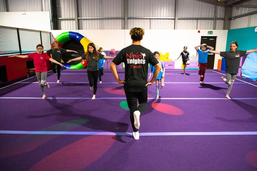 Colourful indoor gymnastics training session with young children balancing on one leg while supervised by a coach in a sports facility equipped with padded mats and play equipment.