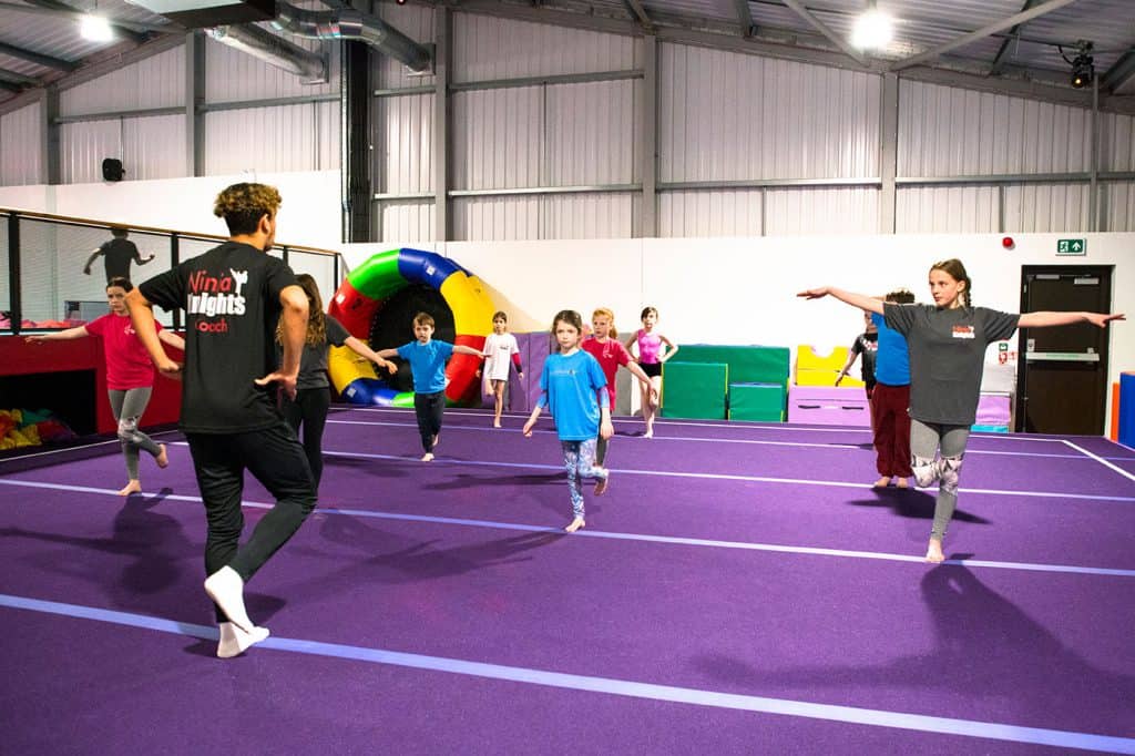 Jumping children participating in a gymnastics class in an indoor sports facility with a purple mat floor.