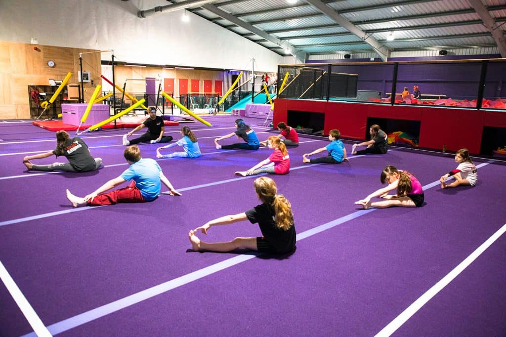 Fun children’s gymnastics class in an indoor trampoline park with purple flooring and gymnastic equipment in the background.