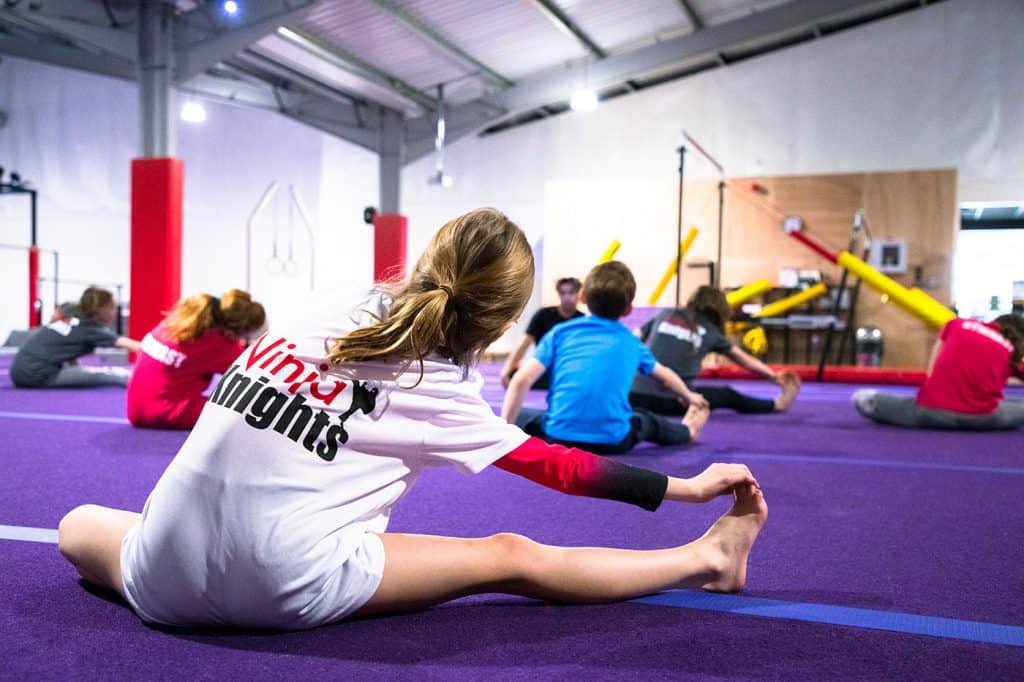 Vinyasa yoga class for kids in indoor gym with colorful equipment and purple mats, focusing on flexibility and stretching exercises. Children are seated on the floor, engaging in a stretching session led by an instructor.