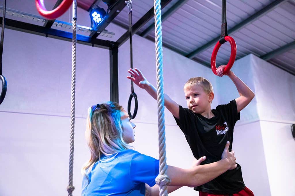 Climbing gym instructor helping a young boy on gymnastic rings during an indoor fitness class. Child training upper body strength with support from a coach in a modern sports facility.