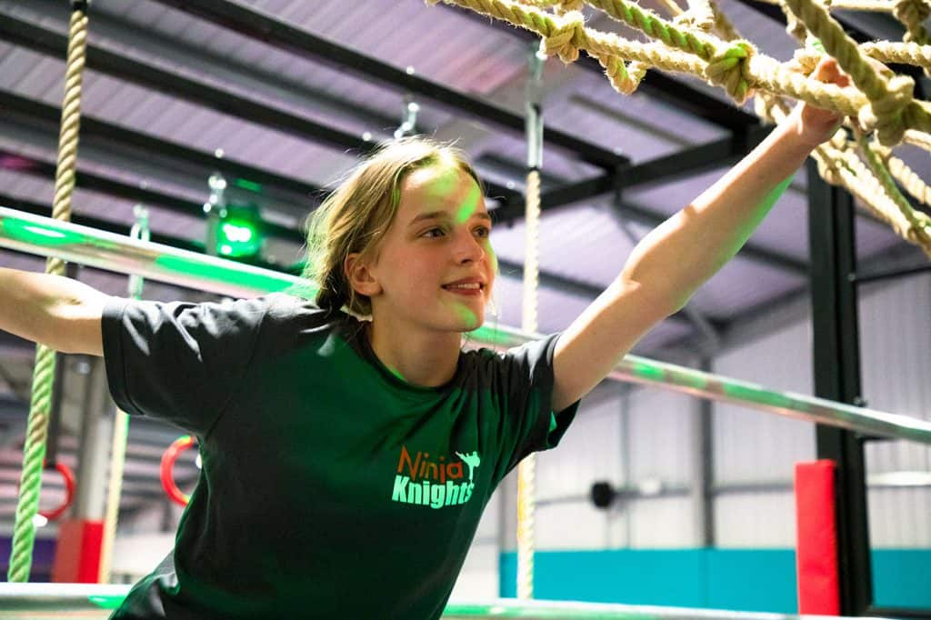 Ninja Knights logo printed on a black T-shirt worn by a young woman extending her arm during an obstacle course race in an indoor activity centre.