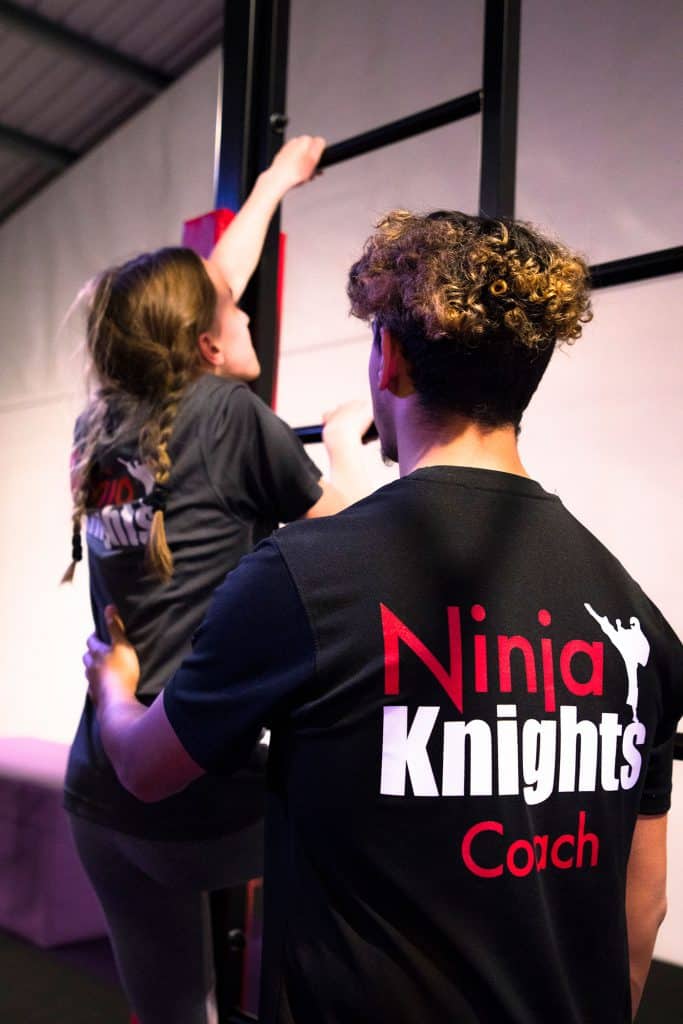 Ninja Knights coach assisting a young girl with a martial arts activity in an indoor gym.