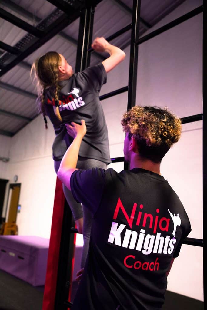 Ninja Knights gym coach assisting a young girl on a climbing wall in a fitness centre. The coach wears a black shirt with the Ninja Knights logo and "Coach" written on the back, providing guidance during the training session.
