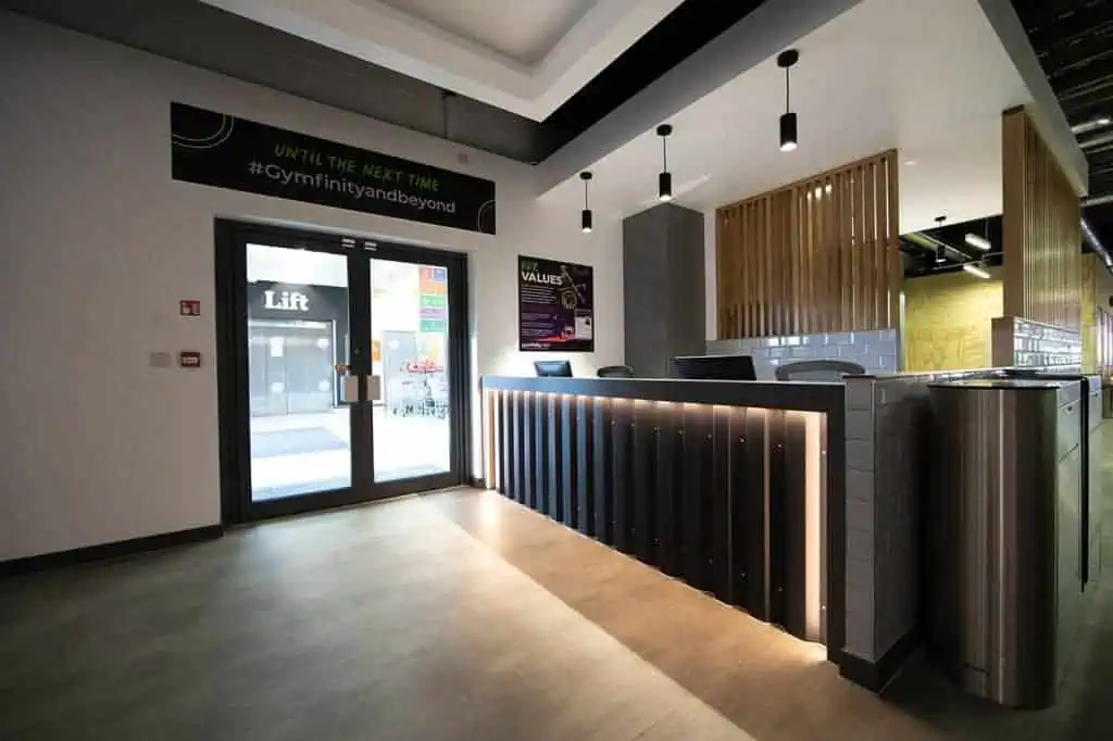 Modern gym reception area with sleek black and metallic accents, LED lighting, wooden partitions, and a glass entrance door, creating a contemporary fitness club atmosphere.